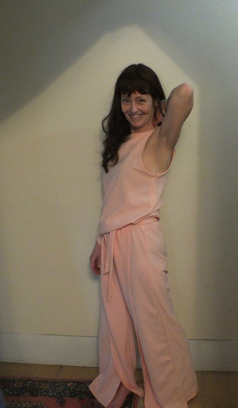 A woman in a jumpsuit that is too big for her standing facing forward against a white wall.